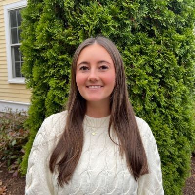 Sarah Hardiman smiles while standing outdoors in front of a lush green shrub, wearing a cream cable-knit sweater, with part of a light-colored building visible in the background.