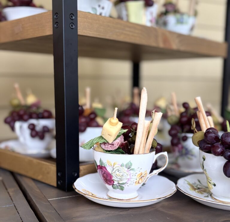 Floral teacups filled with charcuterie, including breadsticks, grapes, cheese, and cured meats, arranged on a wooden surface with more teacups on a shelf in the background.