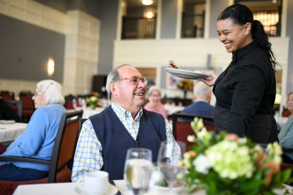 A smiling resident sits at a dining table while a server presents a plated meal, sharing a friendly exchange in a bright, elegant senior living dining room with other residents seated in the background.
