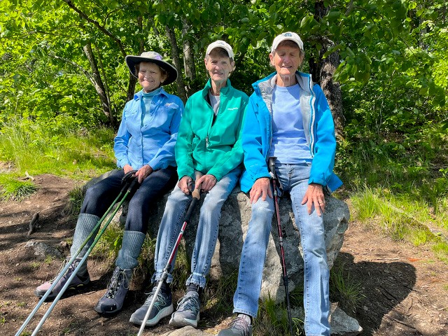 Marilyn Morgan, Jan Regan and Joyce Phillips sit on a rock resting during a hike, with walking poles beside them and greenery in the background. Photo by Judi Foster.