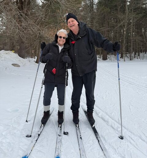 Judi Foster (left) smiles while cross country skiing for the first time alongside a friend on a snowy trail surrounded by trees.