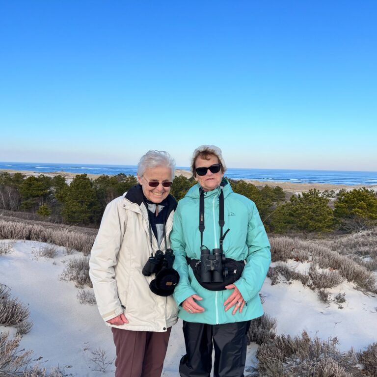 Judi Foster and Marilyn Morgan stand together on a winter beach with binoculars, enjoying an outdoor outing and exploring nature.