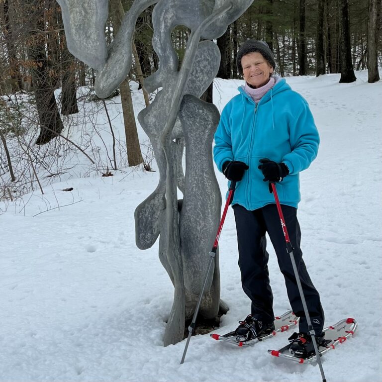 A resident smiles while snowshoeing beside a sculpture at the deCordova Sculpture Park and Museum, enjoying a winter outdoor adventure.