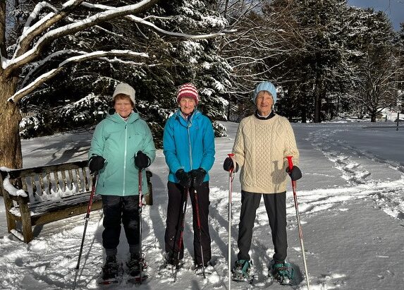 Three women standing outdoors on a snowy path with snowshoes and poles, smiling in winter clothing beside a bench and snow-covered trees on a sunny day.