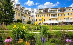Colorful pollinator-friendly flowers bloom in the foreground at The Commons in Lincoln, with native plantings, butterflies and lush garden beds set against the community’s yellow residential buildings under a bright blue sky.