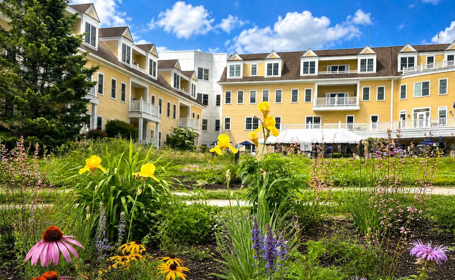 Colorful pollinator-friendly flowers bloom in the foreground at The Commons in Lincoln, with native plantings, butterflies and lush garden beds set against the community’s yellow residential buildings under a bright blue sky.