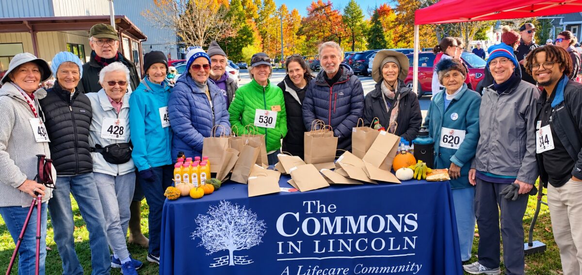 write alt text: Residents and associates of The Commons in Lincoln gather behind a community table at the Scarecrow Classic 5K, celebrating fitness, friendship and community involvement on a colorful fall day.